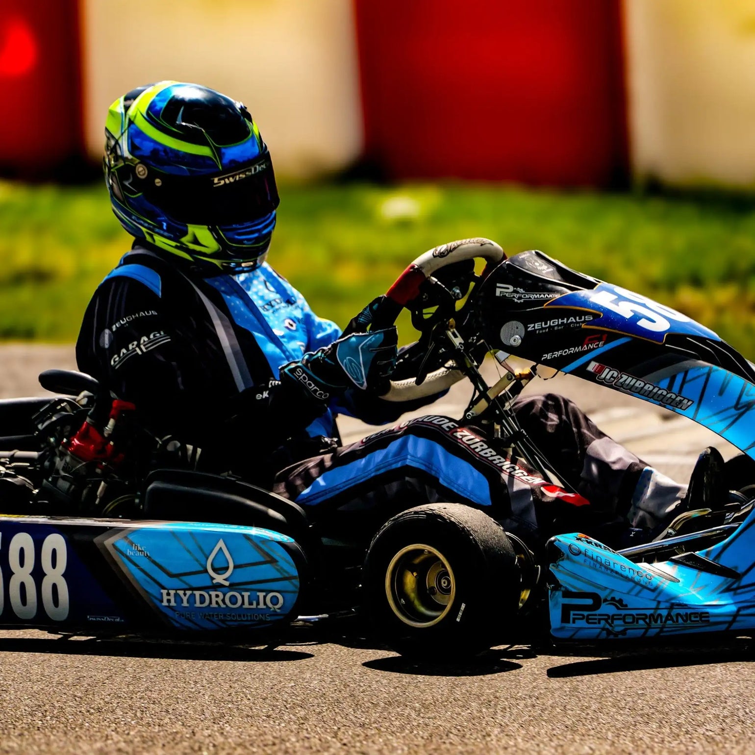 Blauer und schwarzer Go-Kart mit Neongrün-Akzenten, mit Sponsorenlogos wie „hydroliq“ und „zeughaus“, gefahren von einem Fahrer in passender Ausrüstung.