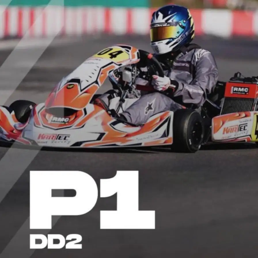 Oranger und weißer Renn-Kart mit schwarzen Reifen, mit Sponsor-Aufklebern mit „’rmc’“ und „’kartec,’“, gefahren von einer Rennfahrerin bzw. einem Rennfahrer in voller Schutzausrüstung.