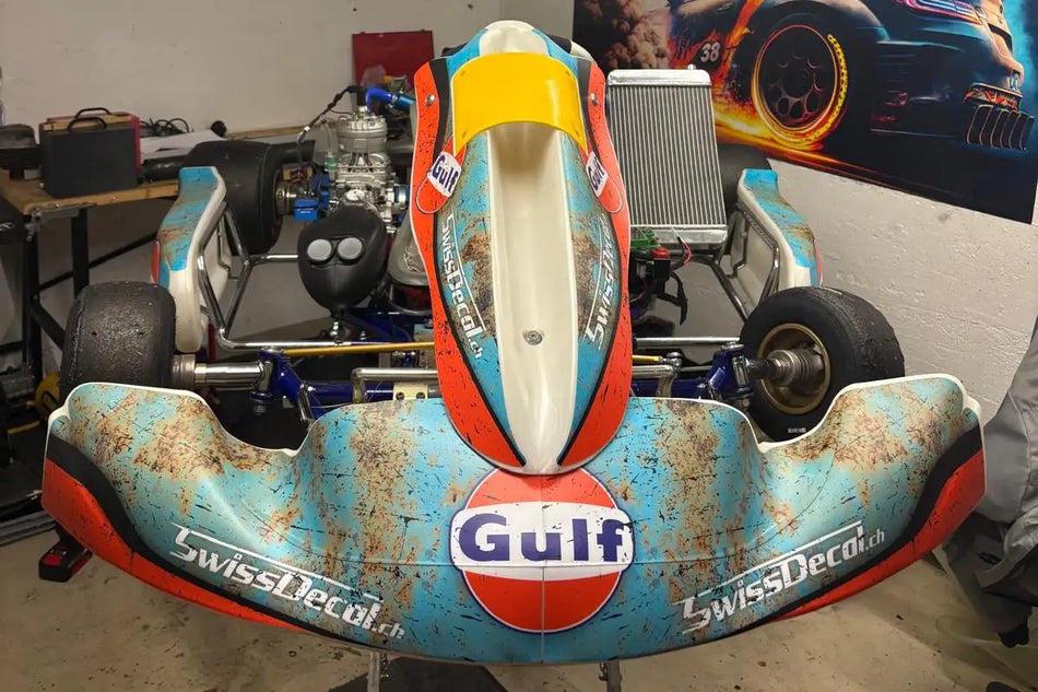 Ein verwitterter türkis- und orangefarbener Go-Kart mit Markenbezug aus der Bucht, mit Rostflecken; mit einem weißen Mittelteil und Aufklebern mit „swissdecal.ch“.
